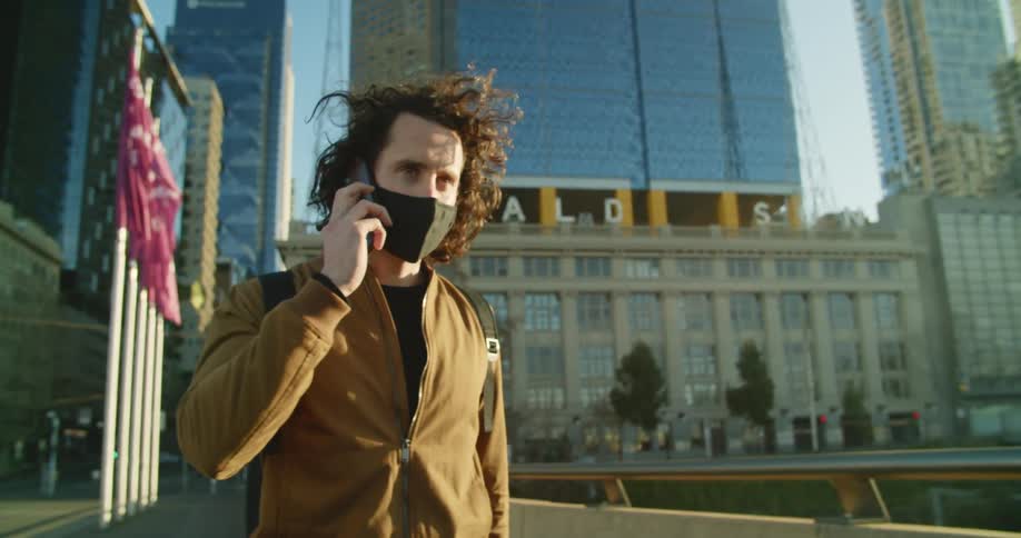 A man in a black medical mask talks on a smartphone while walking down a pathway in a metropolis during the day
