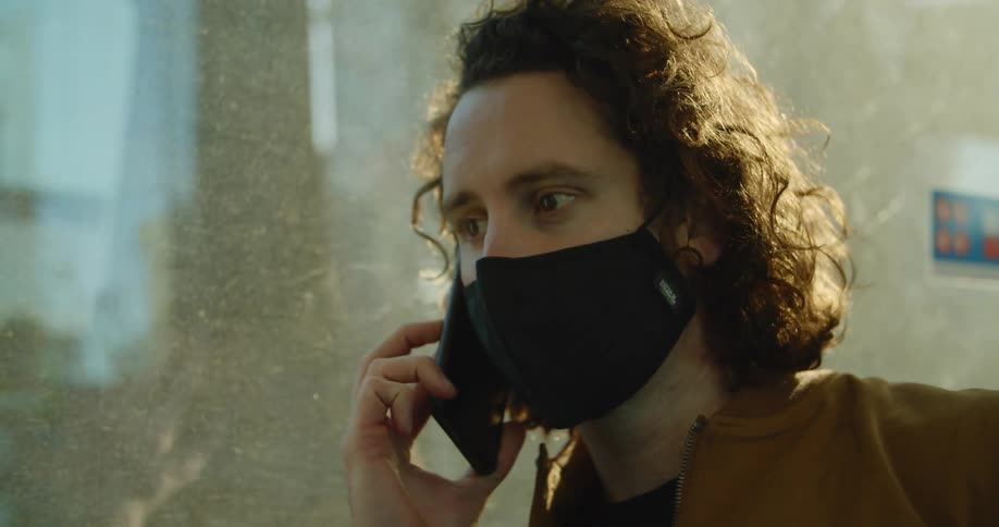 A man with a mask talking on a smartphone on a train next to a window