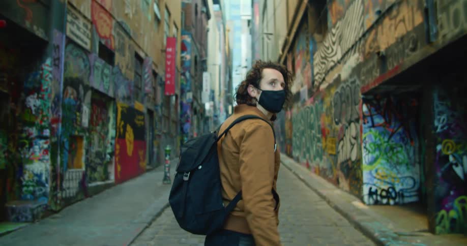 A man in a black medical mask walks through an alleyway with colorful graffiti on every wall