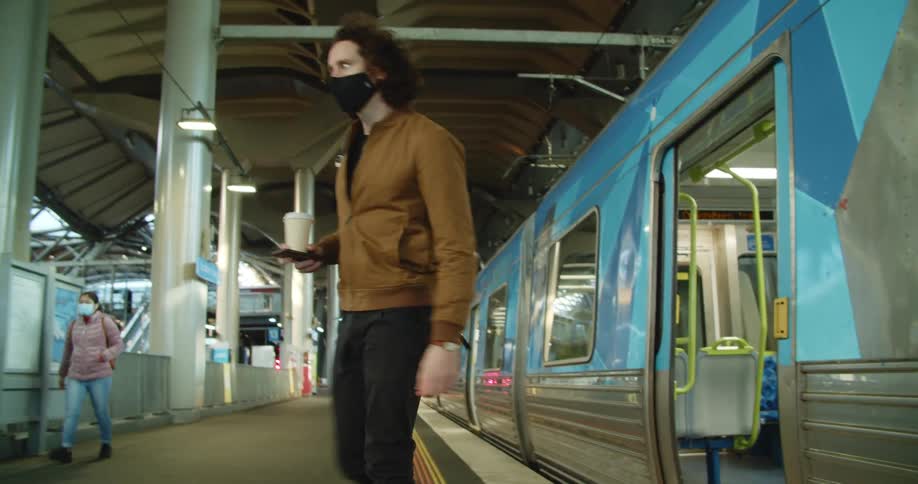 A man in a black medical mask exits a train onto a train platform while balancing a foam cup on his smartphone