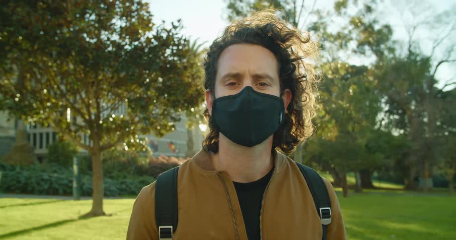Man adjusts face mask standing near lawn in front of building backed by sun