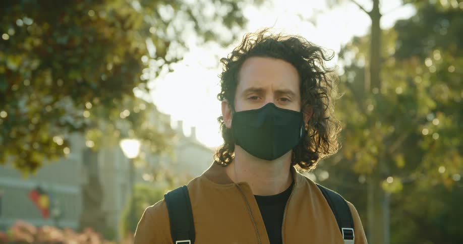 A man puts on a black face mask while standing in a park in a city