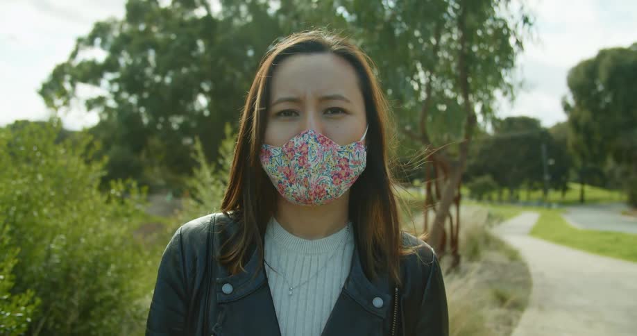 Woman stands near paved path in park wearing face mask as wind rustles branches