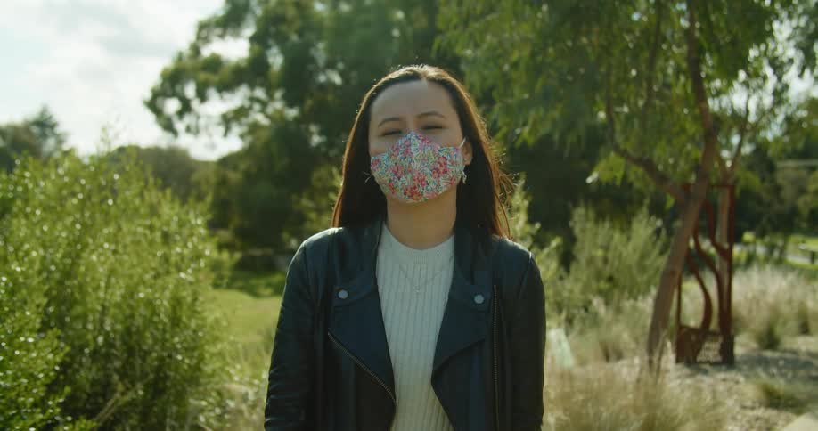 The wind blows the tree and bush branches behind a woman wearing a floral print mask