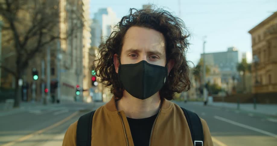 A man stands in the intersection of a multi lane road in a city while wearing a black face mask