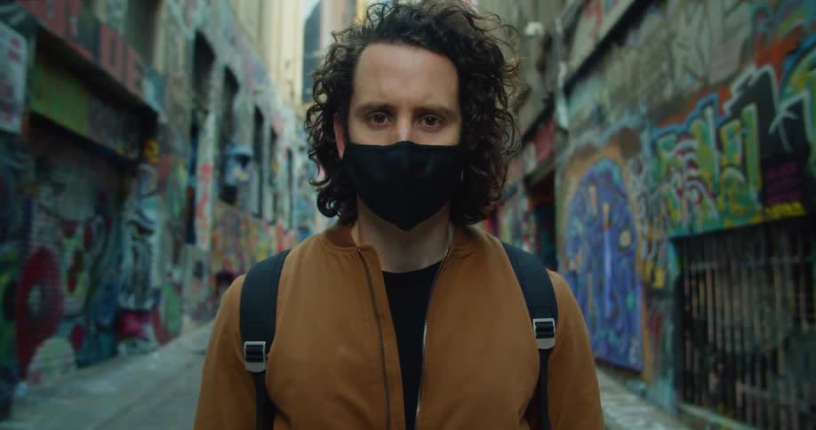 A man wearing a mask stands in a graffiti lined alleyway