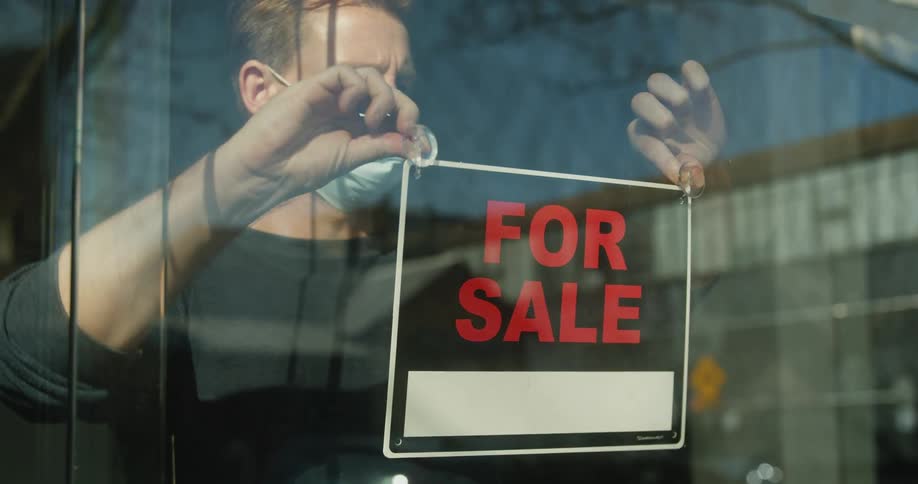 A man wearing a surgical masks places a for sale sign on the inside of a storefront window and walks away