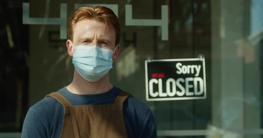 A man wearing a mask stands outside in front of a store with a closed sign in the window