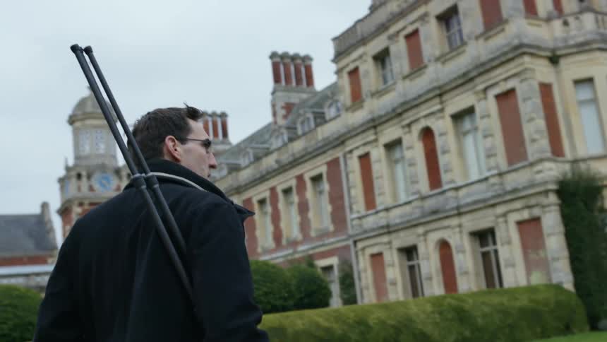 A man walks toward a historic building while carrying a bipod