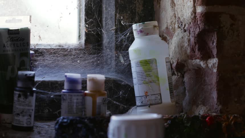 Used bottles of acrylic paint sit in thick cobwebs in a corner by a window