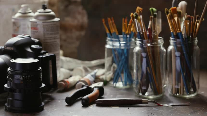 Jars full of paint brushes are on a counter top along with a camera and some spray paint cans