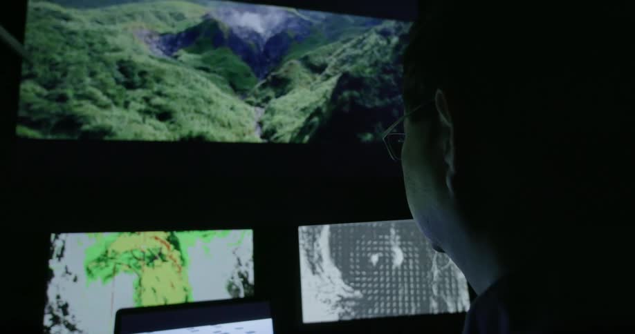 A man sits in front of computer screens and watches them