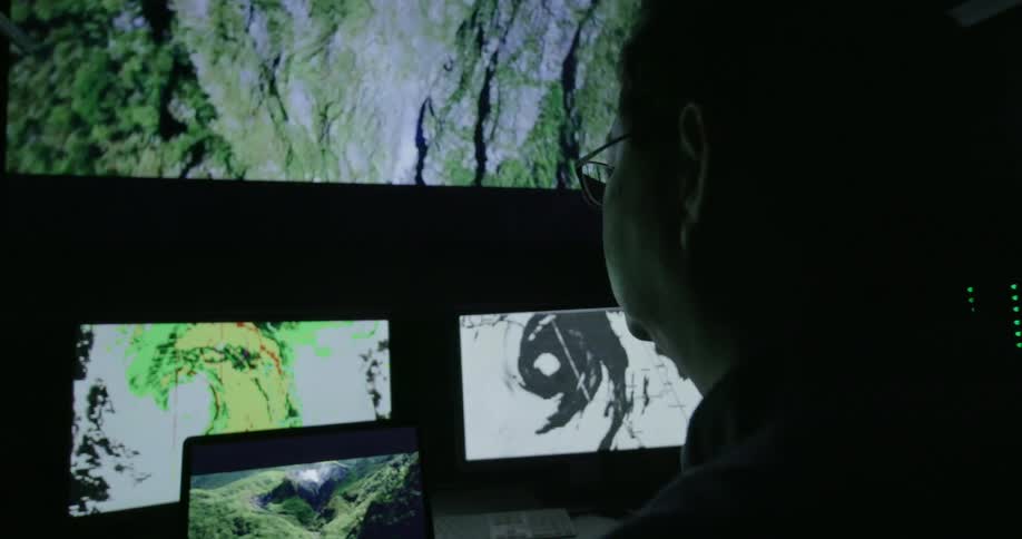 A man sits in front of several computer screens and watches them