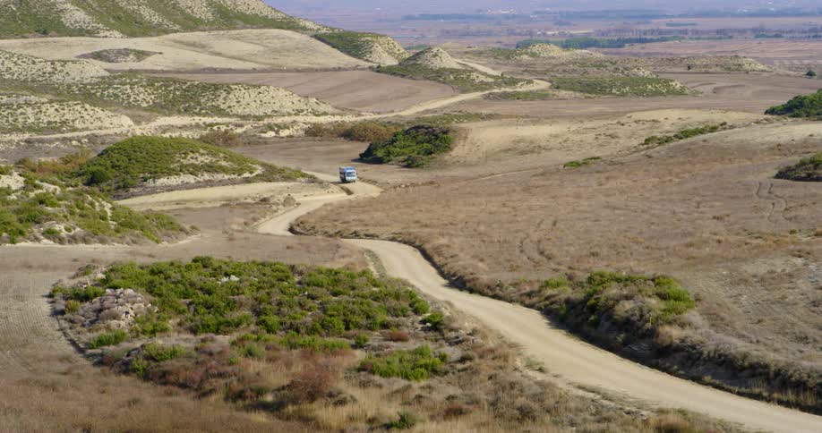 A truck travels on a windy narrow dirt road through a barren landscape dotted with green brush