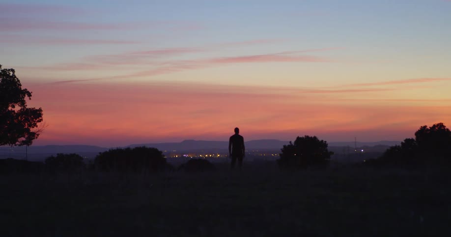 Person stands on a hill looking out at the city below as the sun sets