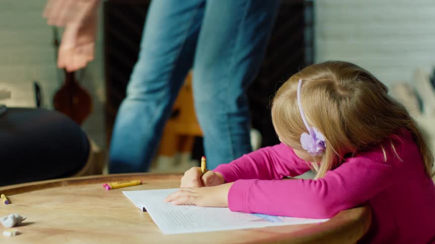 A little girl sits at a table coloring with crayons as her mother picks up a couple of books