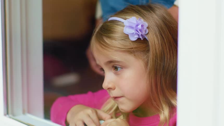 A young girl leans against a window frame and gazes out the window as she speaks to an adult stroking her hair