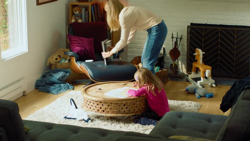 A woman enters a room and collects some books from a chair before stroking the hair of a little girl sitting on a carpet at a table coloring in a coloring book