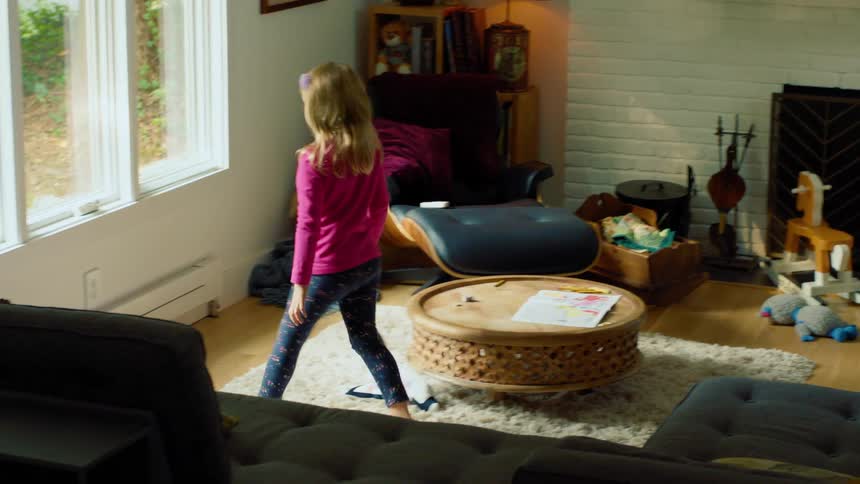 A little girl gets up from coloring at the coffee table and walks over to kneel in front of the window to look outside