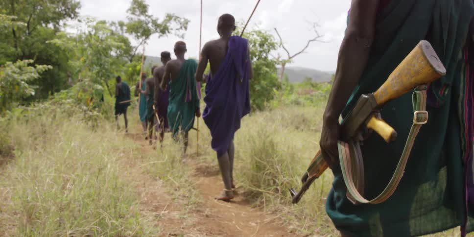 A man with a gun walks down a path with other men holding sticks