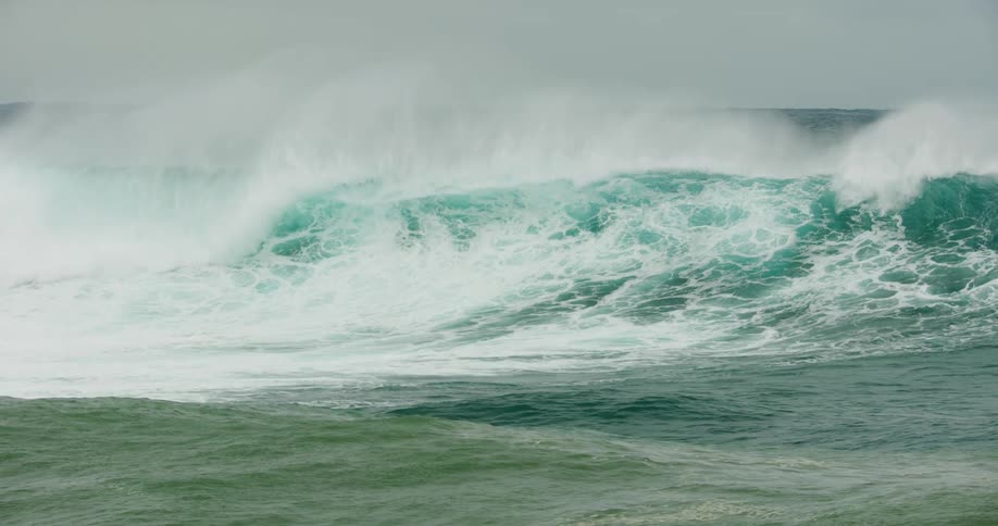 A large ocean wave crashes and sends mist into the air under a pale sky