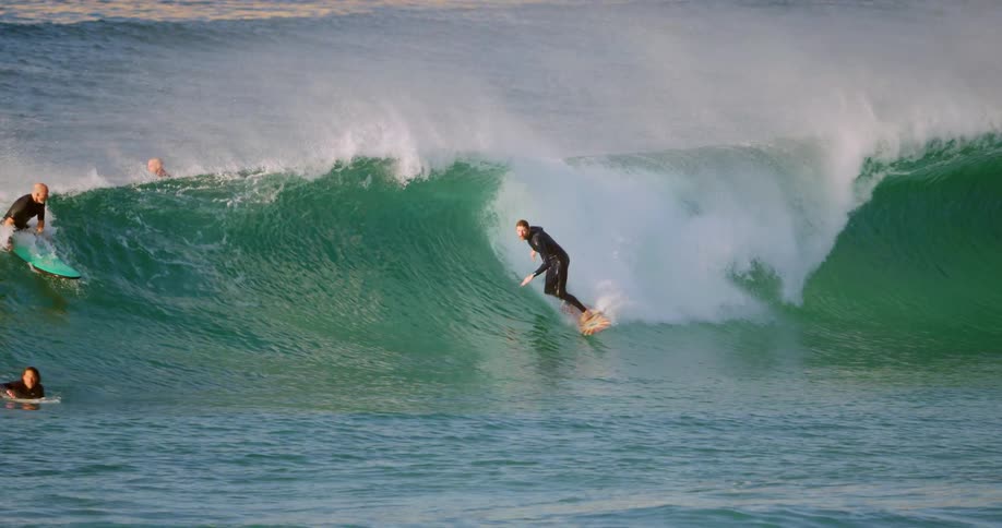 A surfer jumps up on his board and rides a curling breaking wave as other surfers paddle out