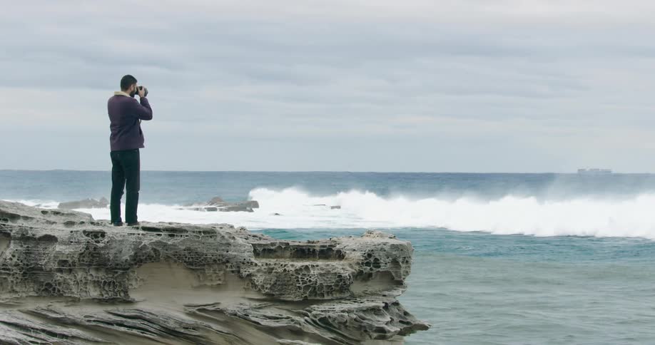 A man stands on an outcropping of rocks to photograph the large waves