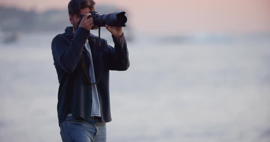 A man standing on shore holds a camera with a long lens photographing the ocean at sunset