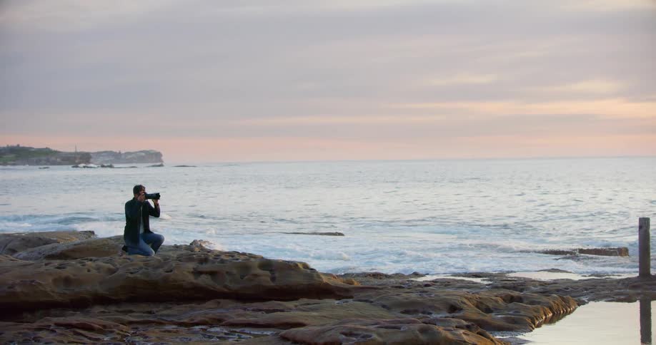 A man kneels on the rocky shore photographing the ocean with a long zoom lens