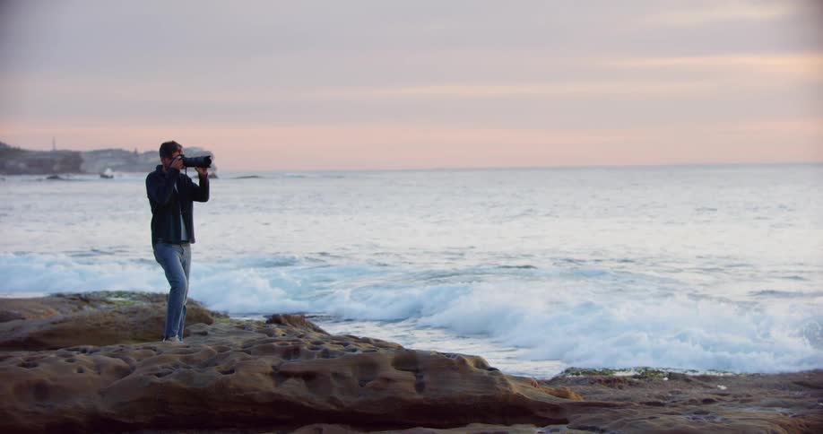 A photographer stands on a rock formation on the coast while taking photographs of the ocean at sunset