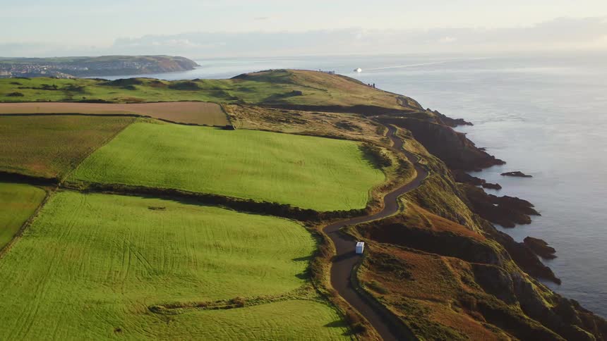 A road near a field follows the curve of a cliff overlooking the ocean