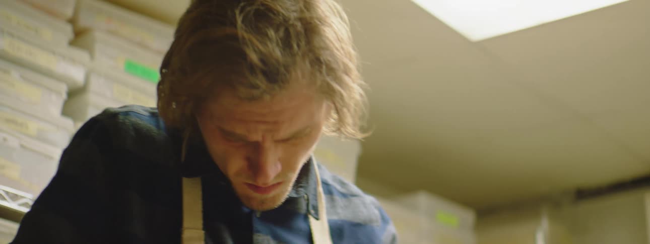 A white man in a gray apron standing in a kitchen cutting a red onion on a wooden cutting board