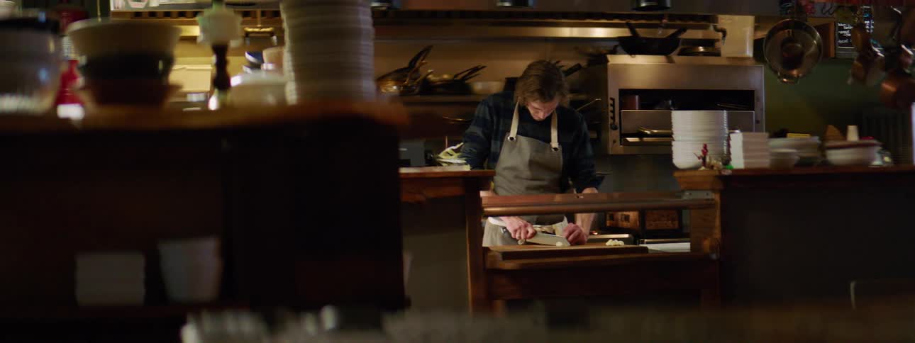 A white man in a gray apron chopping food in a kitchen on a wooden cutting board