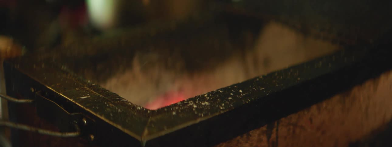 A man bends down and blows on the flames of an indoor smoker grill and places a piece of meat on a skewer across the top
