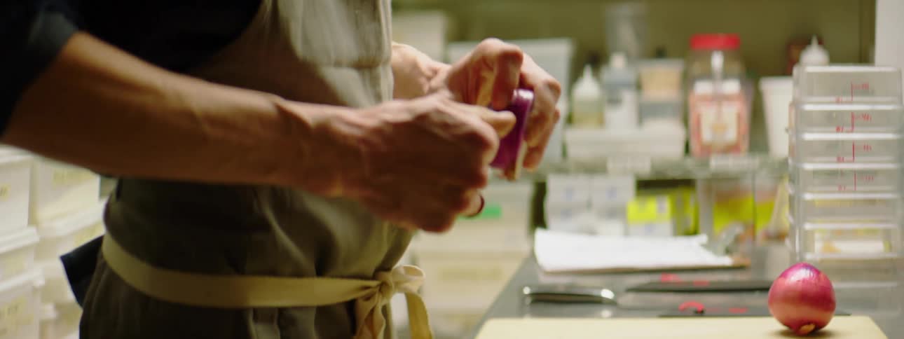 A cook in a gray apron peeling a red onion before sitting in on a wooden cutting board