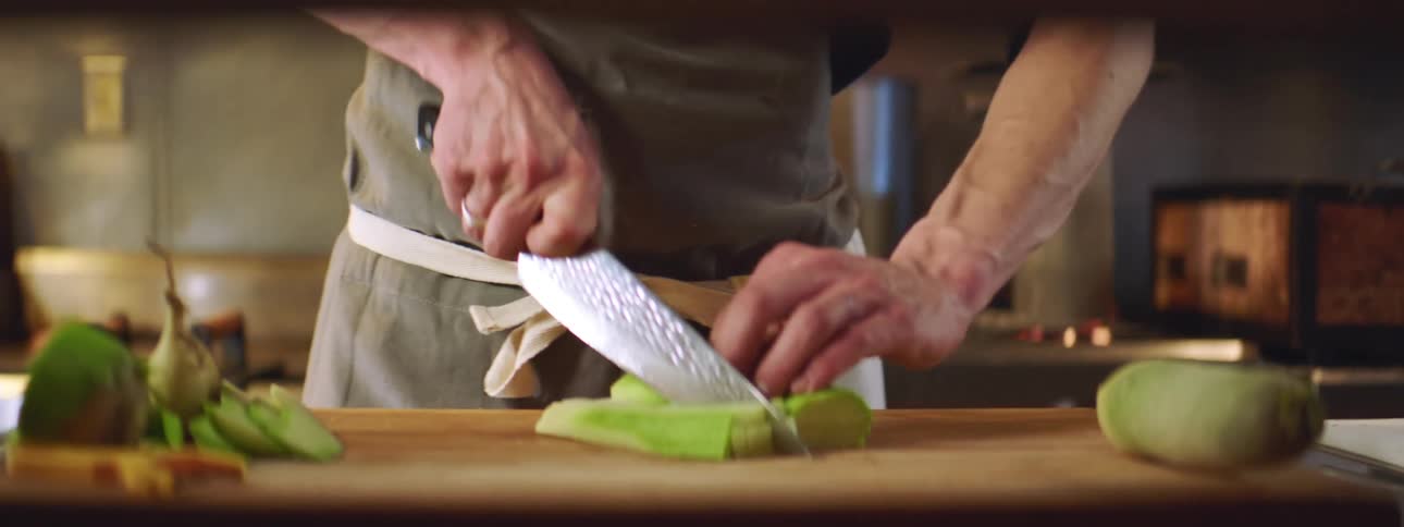 A chef slices through some vegetables while guiding a knife in an up and down motion while being in a home kitchen