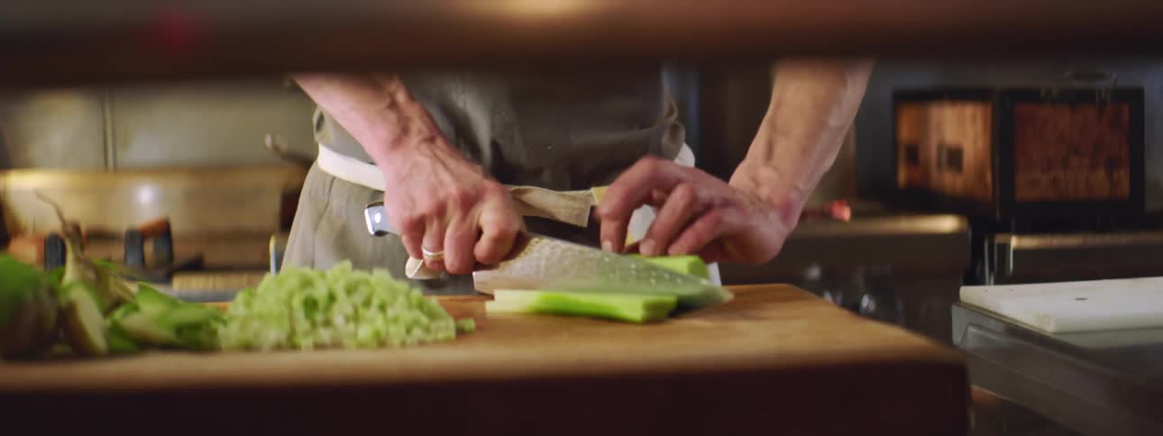 A man in a kitchen wearing a gray apron cutting vegetables on a wooden cutting board