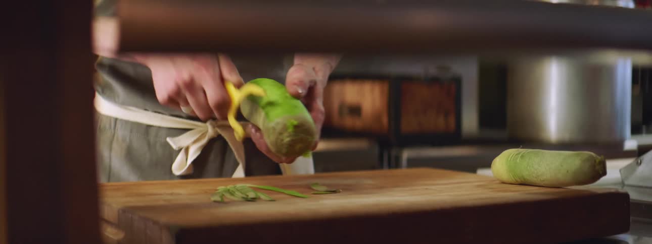 A chef stands in a commercial kitchen peeling a vegetable onto a large cutting board