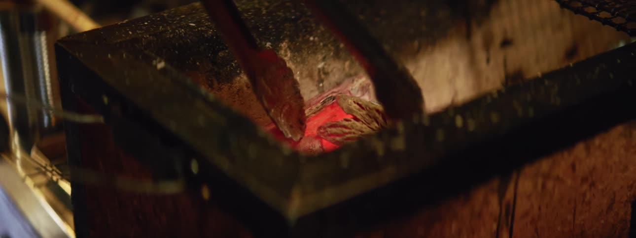 A person using tongs to move pieces of charcoal in a very hot fire pit