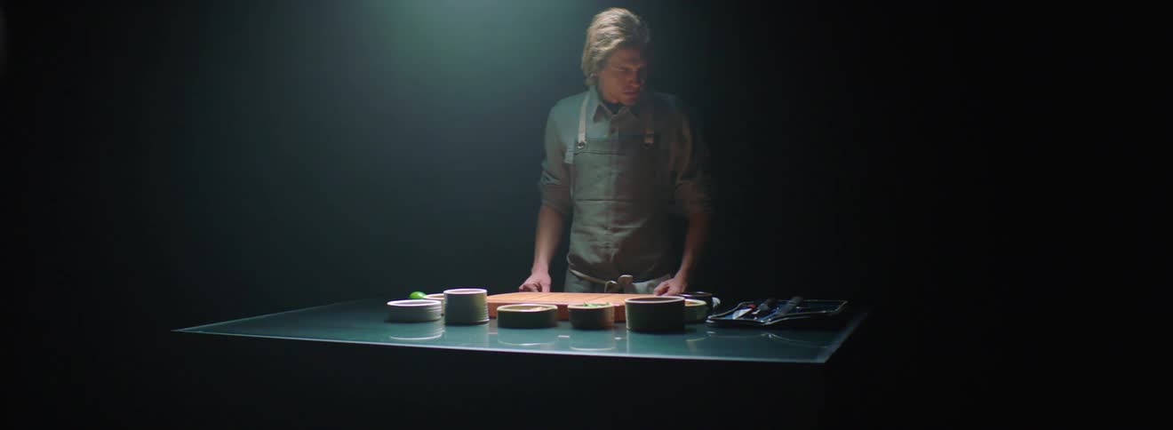 A man wearing an apron walks up to a table lined with cooking utensils and dishes