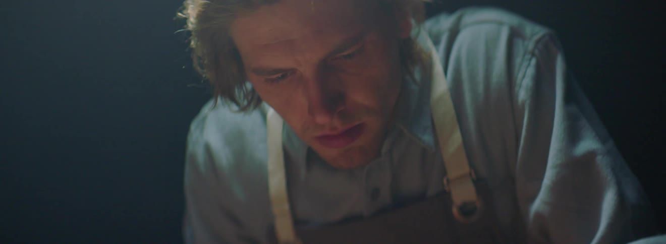 Long hair blonde man wearing apron looks down in the dark background
