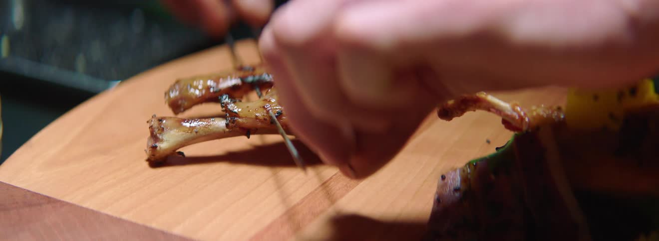 Two skewered pieces of meat on the bone are placed onto a prep table next to an already prepared hors doeuvre