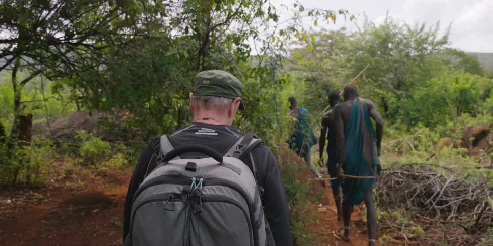 African tribesmen and a caucasian man walking on a path through trees and bushes