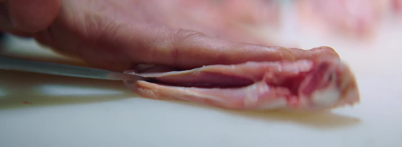 A chef holds down a small piece of meat and slices it open with a sharp knife