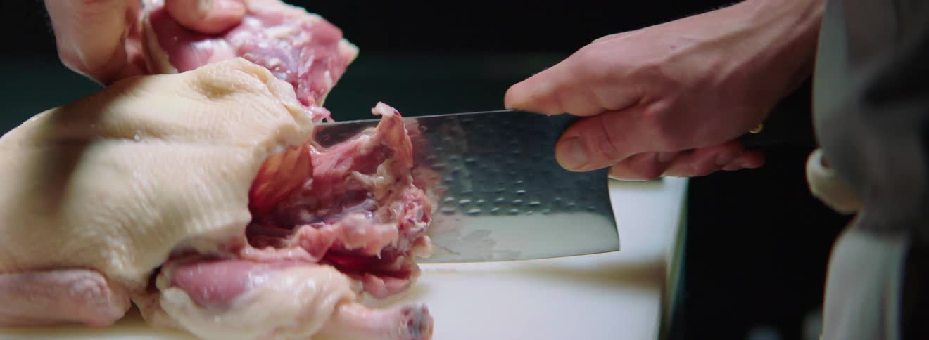 A cook in a dark room cutting chicken with a cleaver