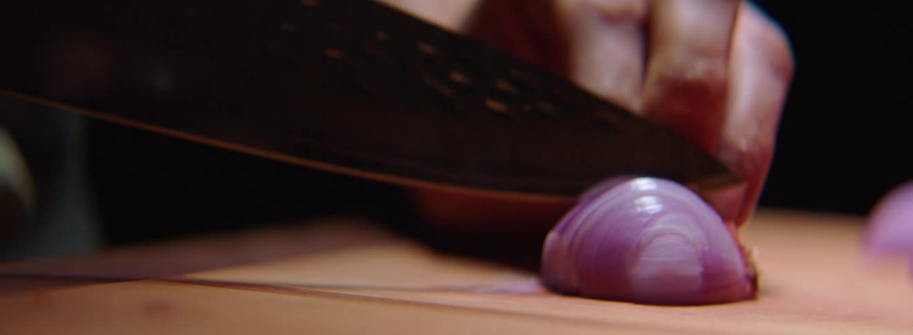 A chef thinly slices a shallot on a cutting board with the tip of a sharp knife