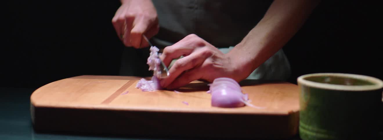 A person in a dark room cutting red onions on a wooden cutting board
