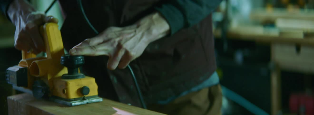 An older white person pushes a sander across a length of wood
