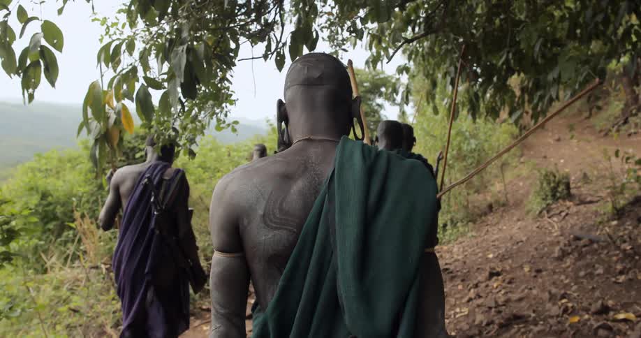 A group of men carrying guns and sticks walk down a dirt path