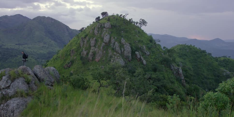 A man stands on a rock near a hill on a cloudy day